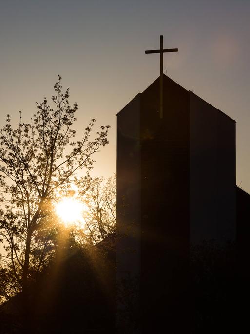 Ein Kirchturm mit Kreuz steht im Gegenlicht der Sonne.