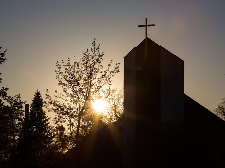 Ein Kirchturm mit Kreuz steht im Gegenlicht der Sonne.