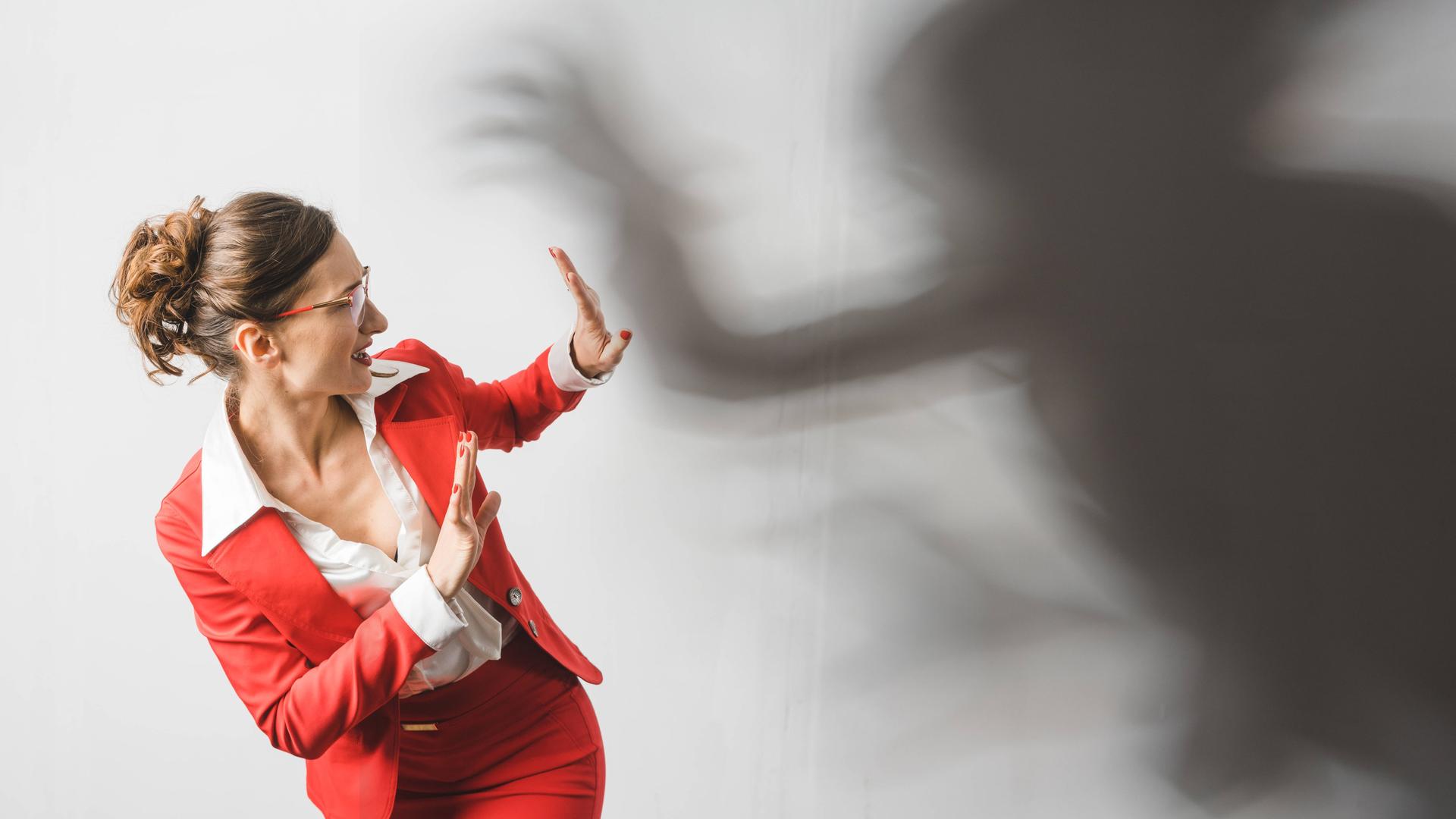 Eine Frau im roten Business-Anzug reagiert abwehrend auf einen schwarzen Schatten, der sich bedrohlich über sie beugt. Symbolbild für Gewalt am Arbeitsplatz