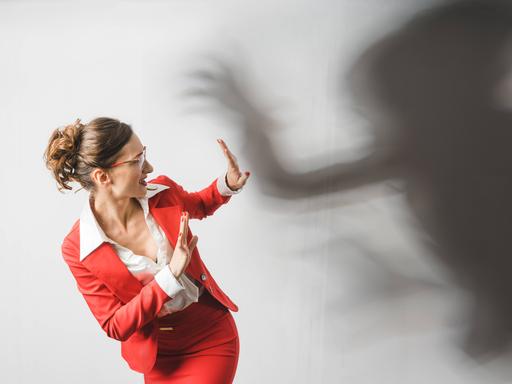 Eine Frau im roten Business-Anzug reagiert abwehrend auf einen schwarzen Schatten, der sich bedrohlich über sie beugt. Symbolbild für Gewalt am Arbeitsplatz Eine Frau im roten Business-Anzug reagiert abwehrend auf einen schwarzen Schatten, der sich bedrohlich über sie beugt. Symbolbild für Gewalt am Arbeitsplatz