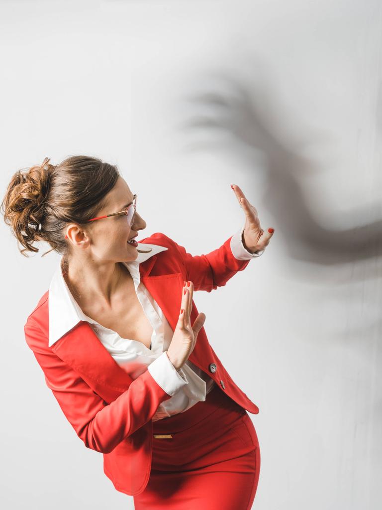 Eine Frau im roten Business-Anzug reagiert abwehrend auf einen schwarzen Schatten, der sich bedrohlich über sie beugt. Symbolbild für Gewalt am Arbeitsplatz Eine Frau im roten Business-Anzug reagiert abwehrend auf einen schwarzen Schatten, der sich bedrohlich über sie beugt. Symbolbild für Gewalt am Arbeitsplatz