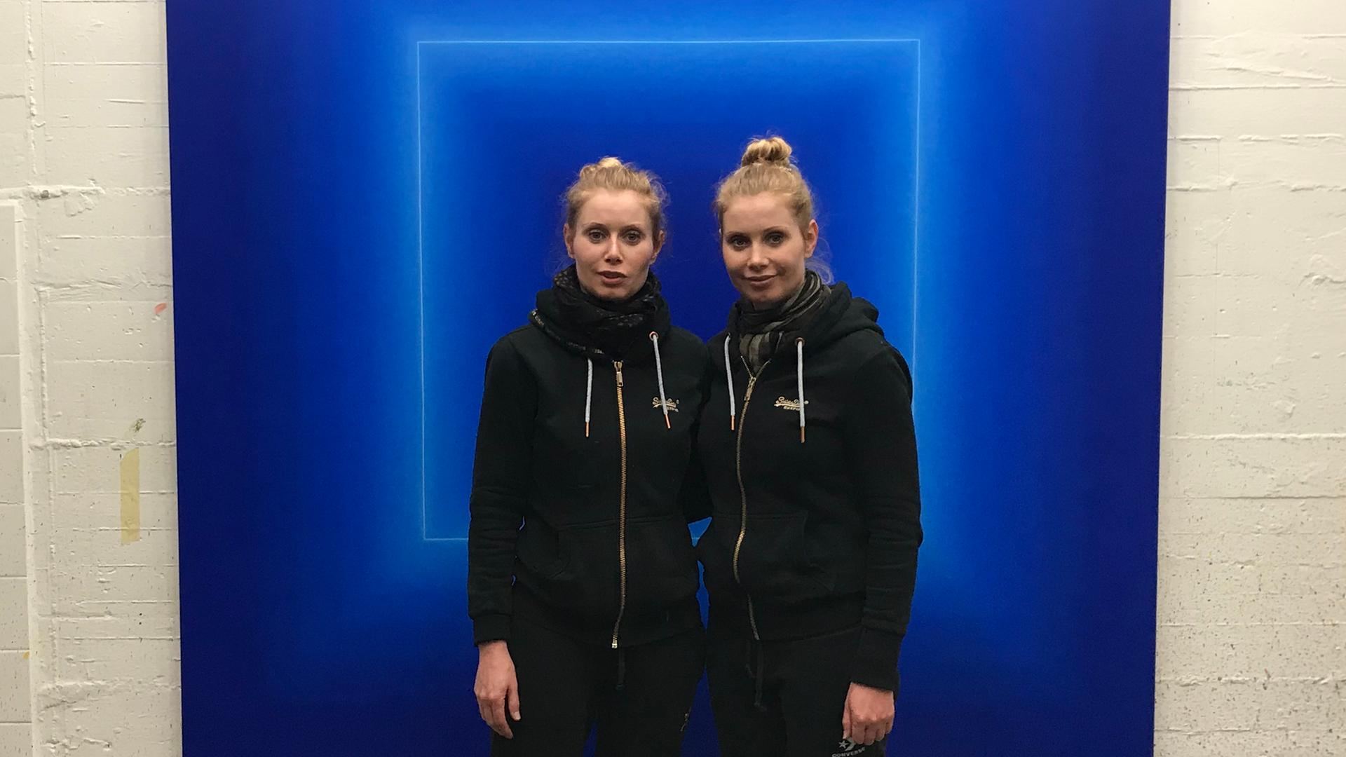 Die beiden Frauen stehen in einem schwarzen Pullover und schwarzen Hosen vor einem großen, blauen Bild. Das Bild hängt in einem großen hallenartigen Raum an einer weißen Wand.