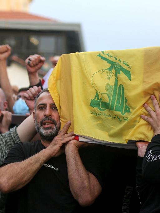 Männer tragen einen Sarg, der mit der libanesischen Flagge und der Flagge der Hisbollah bedeckt ist. Dabei recken sie ihre Fäuste in den Himmel.
