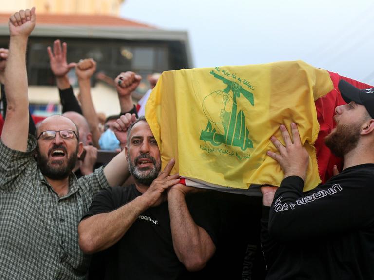 Männer tragen einen Sarg, der mit der libanesischen Flagge und der Flagge der Hisbollah bedeckt ist. Dabei recken sie ihre Fäuste in den Himmel.