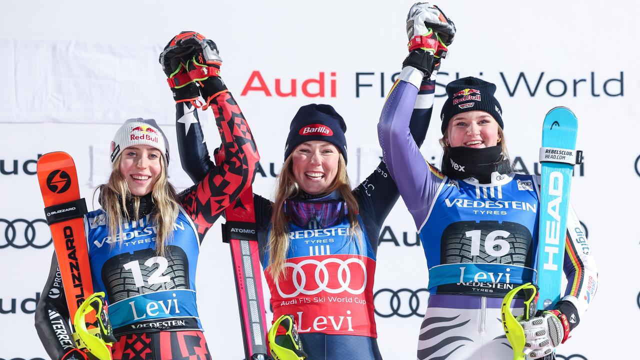 Ski-Weltcup: Lara Colturi (l.), Mikaela Shiffrin (m.) und Emma Aicher (r.) stehen auf dem Podest und halten sich an den Händen, während sie ihre Arme in die Höhe strecken. Alle drei lachen und freuen sich über ihren Erfolg.