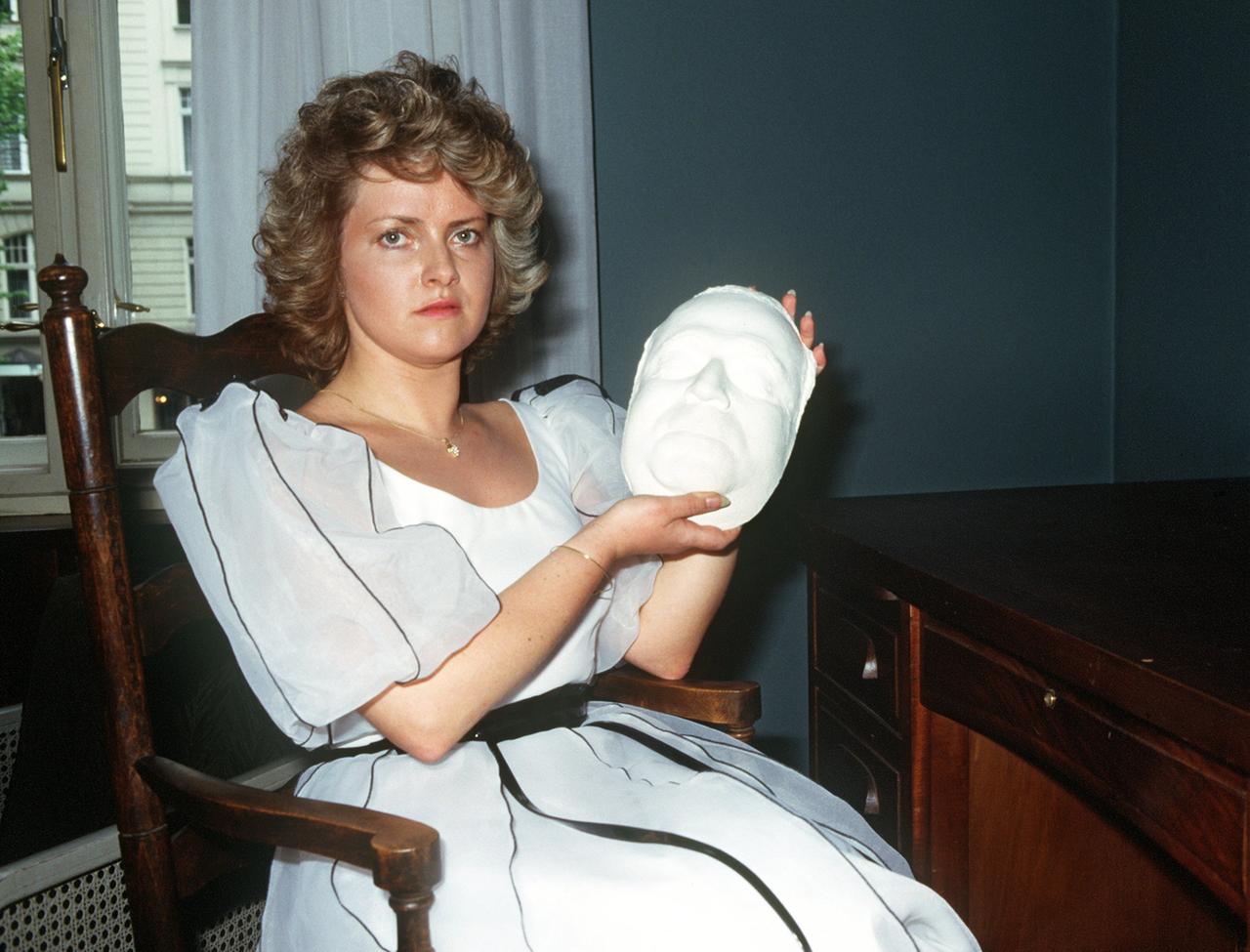 Eine junge Frau sitzt auf einem Stuhl und hält eine Ausfertigung der Totenmaske von Kurt Tucholsky in der Hand. 