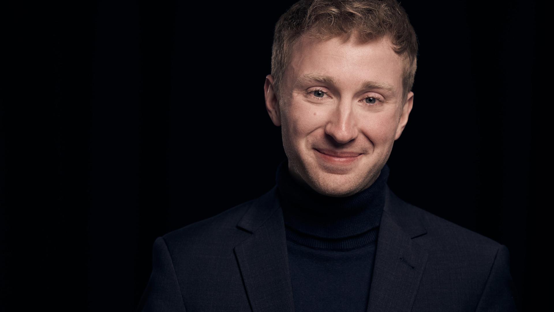 Ein junger Mann mit rotblondem, kurzem Haar blickt lächelnd in die Kamera. Er trägt einen schwarzen Anzug und steht in einem ganz schwarzen Raum, das einzig Helle ist sein Gesicht.