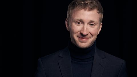 Ein junger Mann mit rotblondem, kurzem Haar blickt lächelnd in die Kamera. Er trägt einen schwarzen Anzug und steht in einem ganz schwarzen Raum, das einzig Helle ist sein Gesicht.