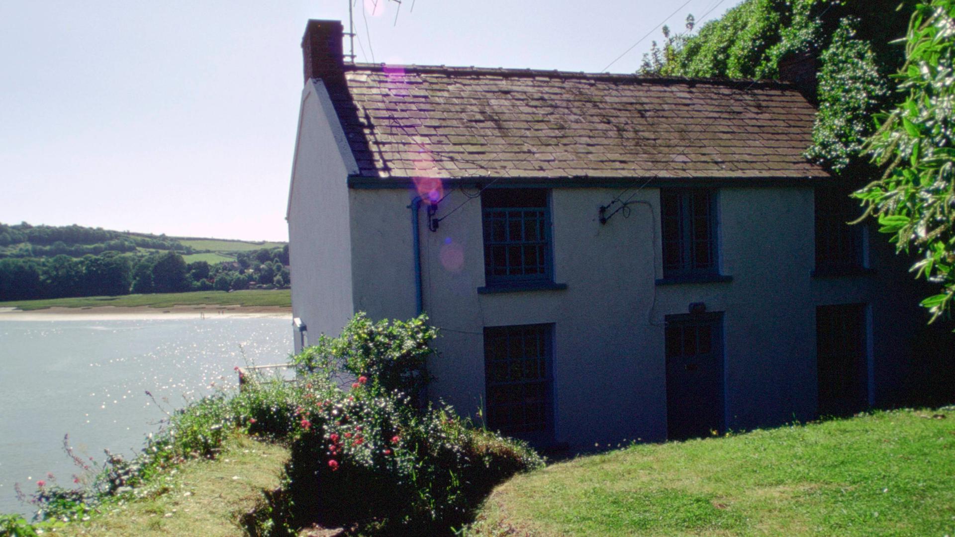 Ein Bootshaus mit einer Antenne drauf steht am Wasser. Durch einen Rasen bewachsenen Garten führt ein Weg zum Haus.