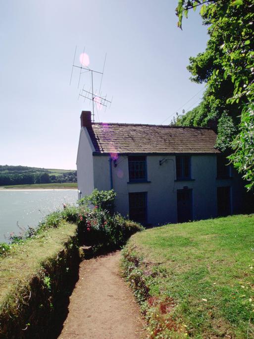 Ein Bootshaus mit einer Antenne drauf steht am Wasser. Durch einen Rasen bewachsenen Garten führt ein Weg zum Haus. Ein Bootshaus mit einer Antenne drauf steht am Wasser. Durch einen Rasen bewachsenen Garten führt ein Weg zum Haus.