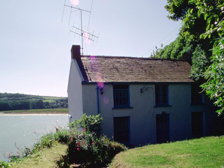 Ein Bootshaus mit einer Antenne drauf steht am Wasser. Durch einen Rasen bewachsenen Garten führt ein Weg zum Haus. Ein Bootshaus mit einer Antenne drauf steht am Wasser. Durch einen Rasen bewachsenen Garten führt ein Weg zum Haus.
