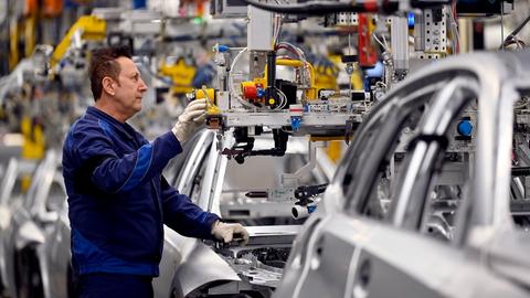 Ein Arbeiter eines Automobilherstellers steht in einer Fabrik an einer Fertigungsstraße, an der halbfertige Wagen durchlaufen. 