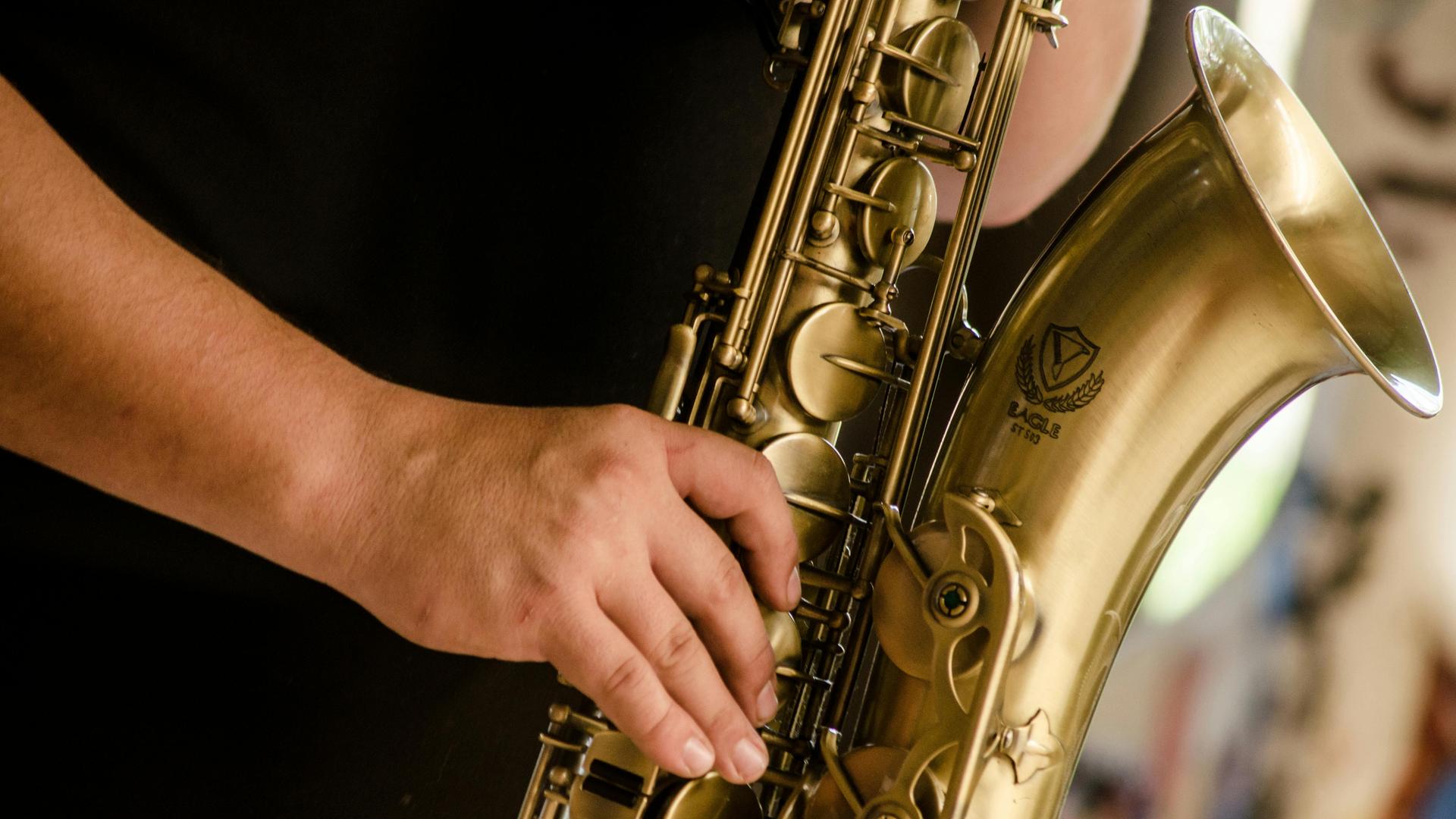 Detailblick auf eine Männerhand, die ein Tenorsaxopon spielt. Man sieht die großen Klappen und den Trichter des Instrumentes.