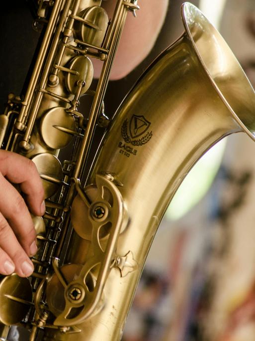 Detailblick auf eine Männerhand, die ein Tenorsaxopon spielt. Man sieht die großen Klappen und den Trichter des Instrumentes.