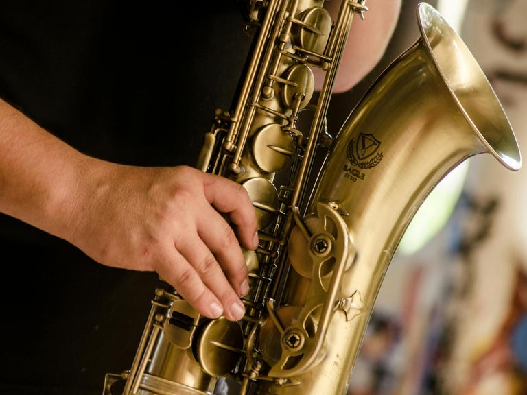 Detailblick auf eine Männerhand, die ein Tenorsaxopon spielt. Man sieht die großen Klappen und den Trichter des Instrumentes.