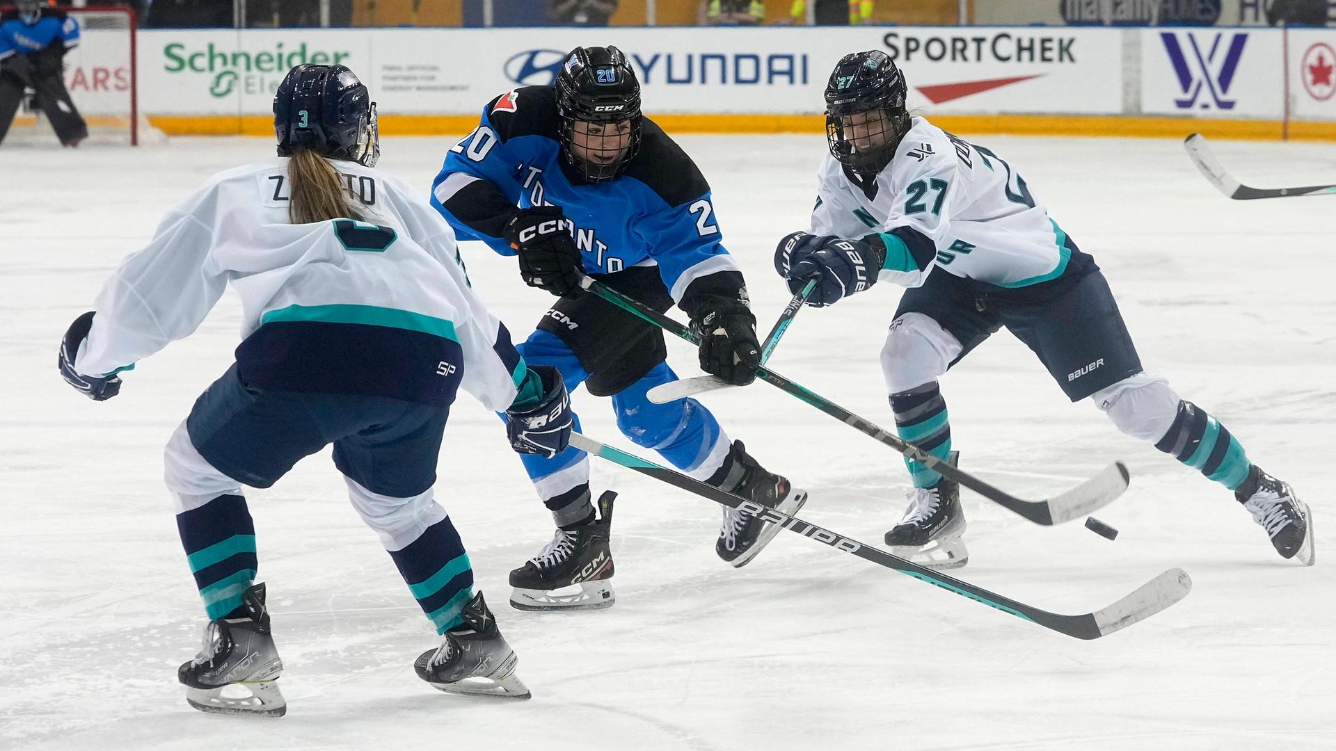 Zwei Eishockey-Spielerinnen vom Team aus New York in weißen Trikots versuchen einer Spielerin aus Toronto im blauen Trikot den Puck abzunehmen.