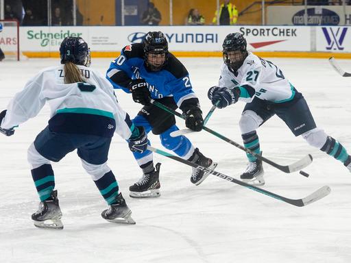 Zwei Eishockey-Spielerinnen vom Team aus New York in weißen Trikots versuchen einer Spielerin aus Toronto im blauen Trikot den Puck abzunehmen.