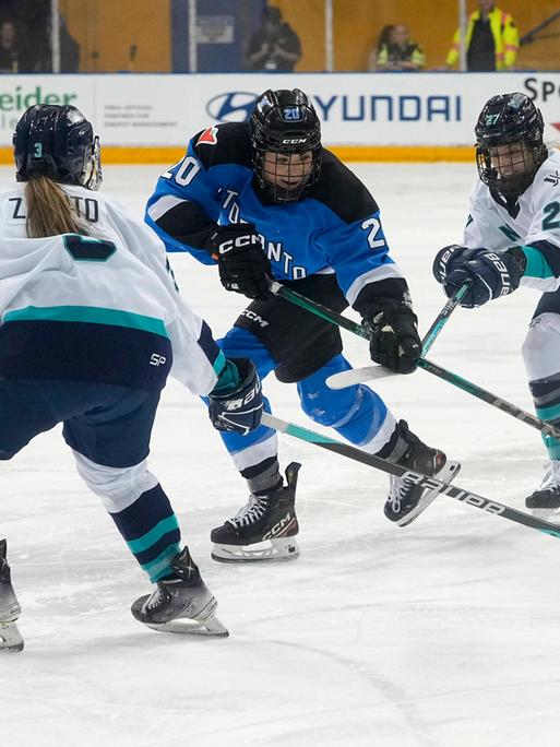 Zwei Eishockey-Spielerinnen vom Team aus New York in weißen Trikots versuchen einer Spielerin aus Toronto im blauen Trikot den Puck abzunehmen.