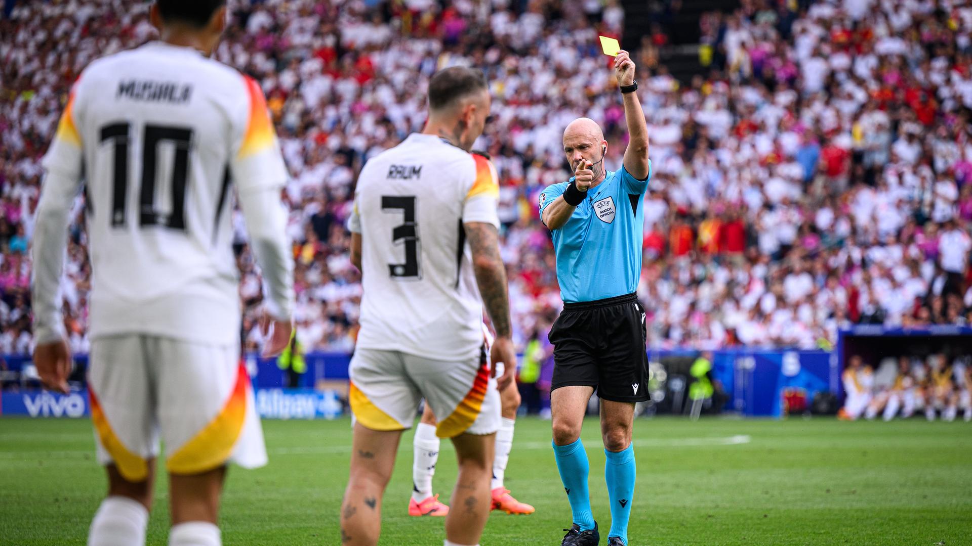 Anthony Taylor zeigt die gelbe Karte. Im Hintergrund sind die Fans im Stadion zu sehen, im Vordergrund, in der Unschärfe, zwei deutsche Spieler