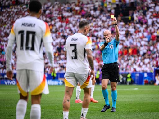 Anthony Taylor zeigt die gelbe Karte. Im Hintergrund sind die Fans im Stadion zu sehen, im Vordergrund, in der Unschärfe, zwei deutsche Spieler