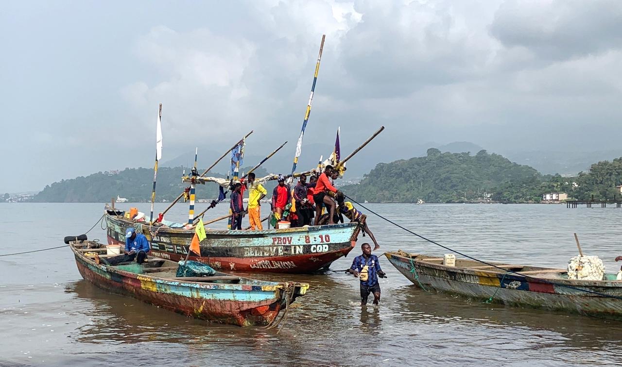 Ein altes Boot mit mehreren jungen Männern landet am Strand. Einer der Fischer ist schon ins Wasser gesprungen. Im Hintergrund sind Inseln und Berge zu sehen.