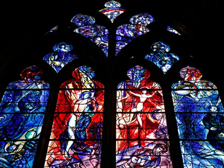 In der Kathedrale von Metz ist ein farbiges Glasfenster zu sehen. Das Fenster ist in vier große und mehrere kleine Teile unterteilt. Auf den großen Fenstern sind fragmentarisch Personen in Bewegung zu erkennen.