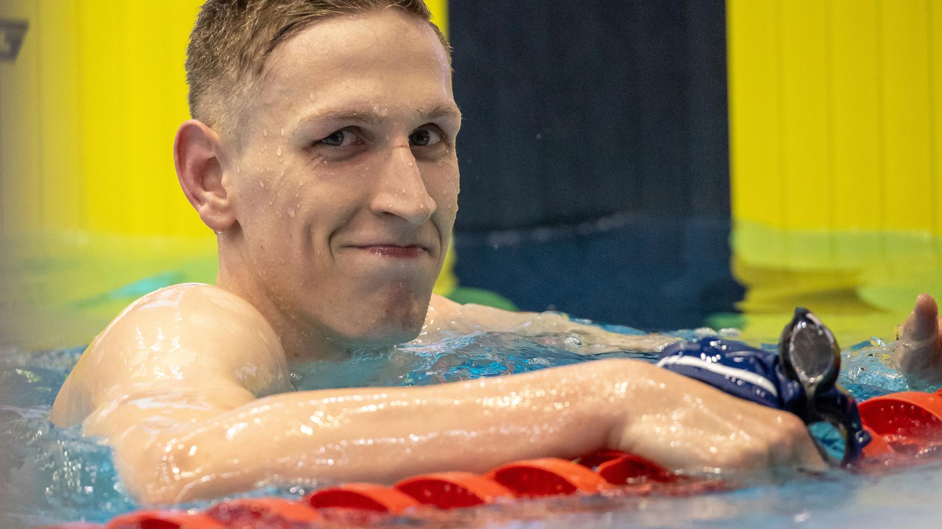 Lukas Märtens im Schwimmbecken. Er hält sich an der Wettkampfleine fest und grinst verschmitzt in die Kamera.