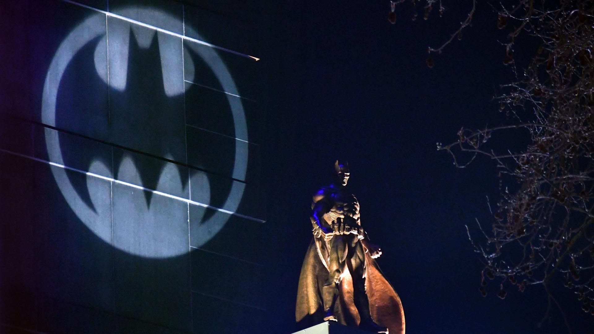 Auf einem Hochhaus wird das Symbol von Batman angestrahlt, davor steht eine große Batman-Figur auf einem anderen Häuserdach.