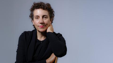 Eine Frau mit dunklem, kurz gelocktem Haar trägt dunkle Kleidung und blickt freundlich in die Kamera. Sie sitzt vor taubenblauem Hintergrund seitlich auf einem Stuhl.