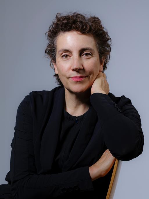 Eine Frau mit dunklem, kurz gelocktem Haar trägt dunkle Kleidung und blickt freundlich in die Kamera. Sie sitzt vor taubenblauem Hintergrund seitlich auf einem Stuhl.