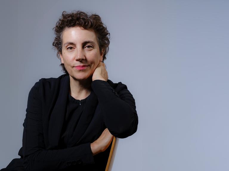 Eine Frau mit dunklem, kurz gelocktem Haar trägt dunkle Kleidung und blickt freundlich in die Kamera. Sie sitzt vor taubenblauem Hintergrund seitlich auf einem Stuhl.