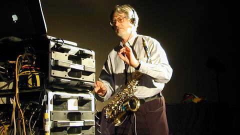 Bruno Spoerri steht mit einem Saxophon um den Hals in einem Raum, in dem Studio-Ausrüstung steht. Er trägt einen Bart und hat Kopfhörer auf, die Hände hält er wie ein Dirigent.