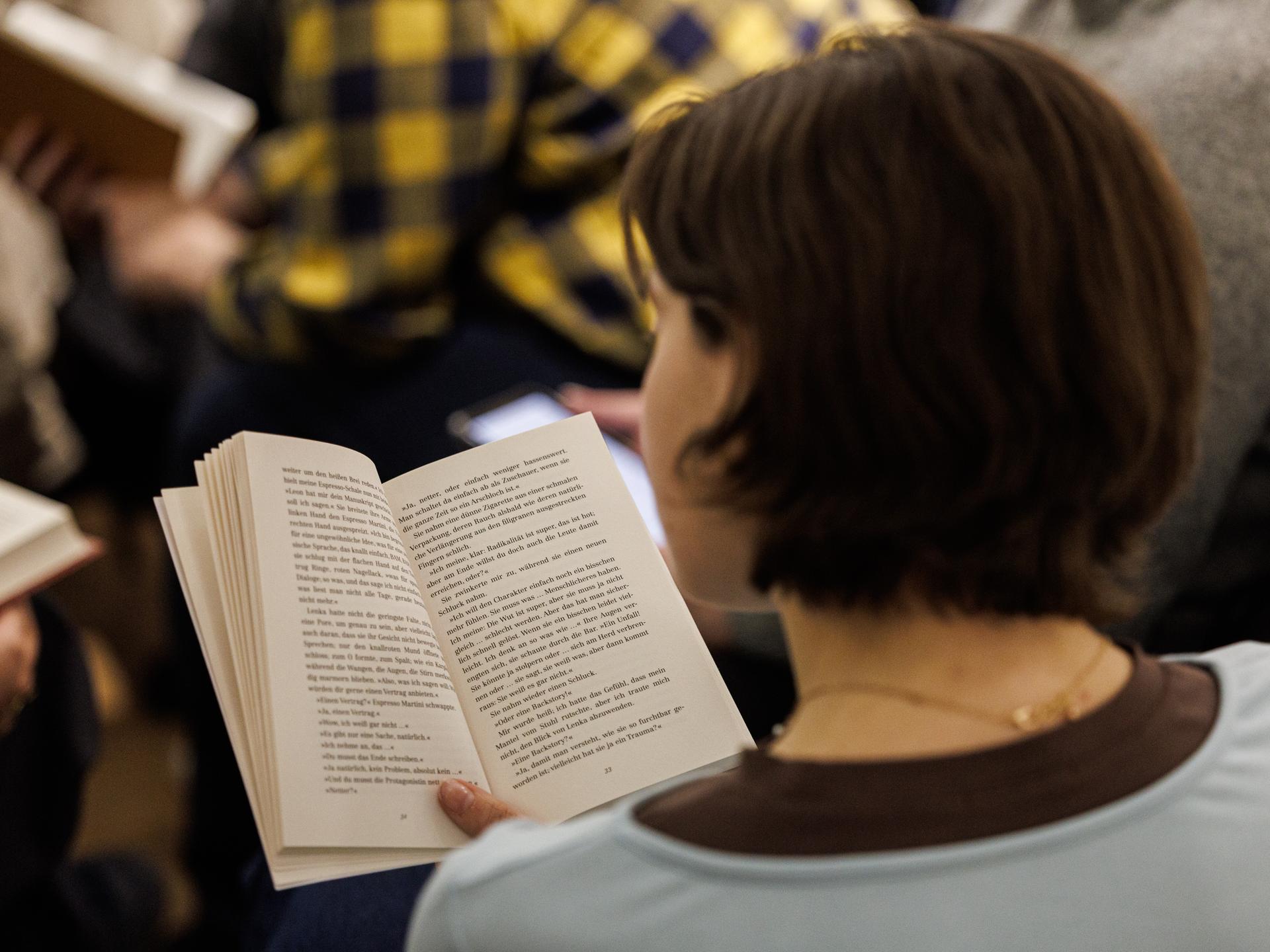 Eine Frau mit dunklen Haaren ist von hinten zu sehen. Sie hält ein Buch in der Hand und liest. 