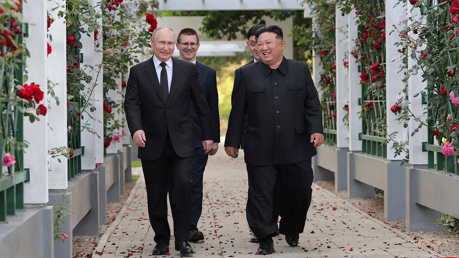 Wladimir Putin und Kim Jong Un läufen unter einer weißen Pergola, die mit roten Rosen umrankt ist und lachen.