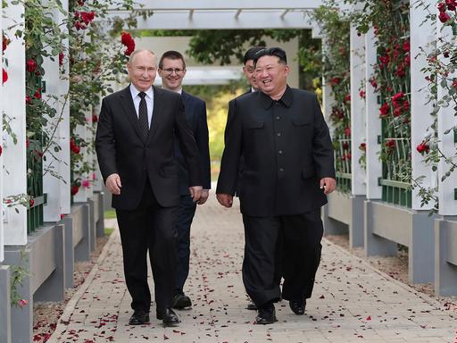 Wladimir Putin und Kim Jong Un läufen unter einer weißen Pergola, die mit roten Rosen umrankt ist und lachen.