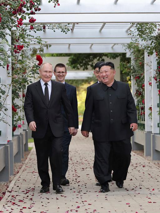 Wladimir Putin und Kim Jong Un läufen unter einer weißen Pergola, die mit roten Rosen umrankt ist und lachen.