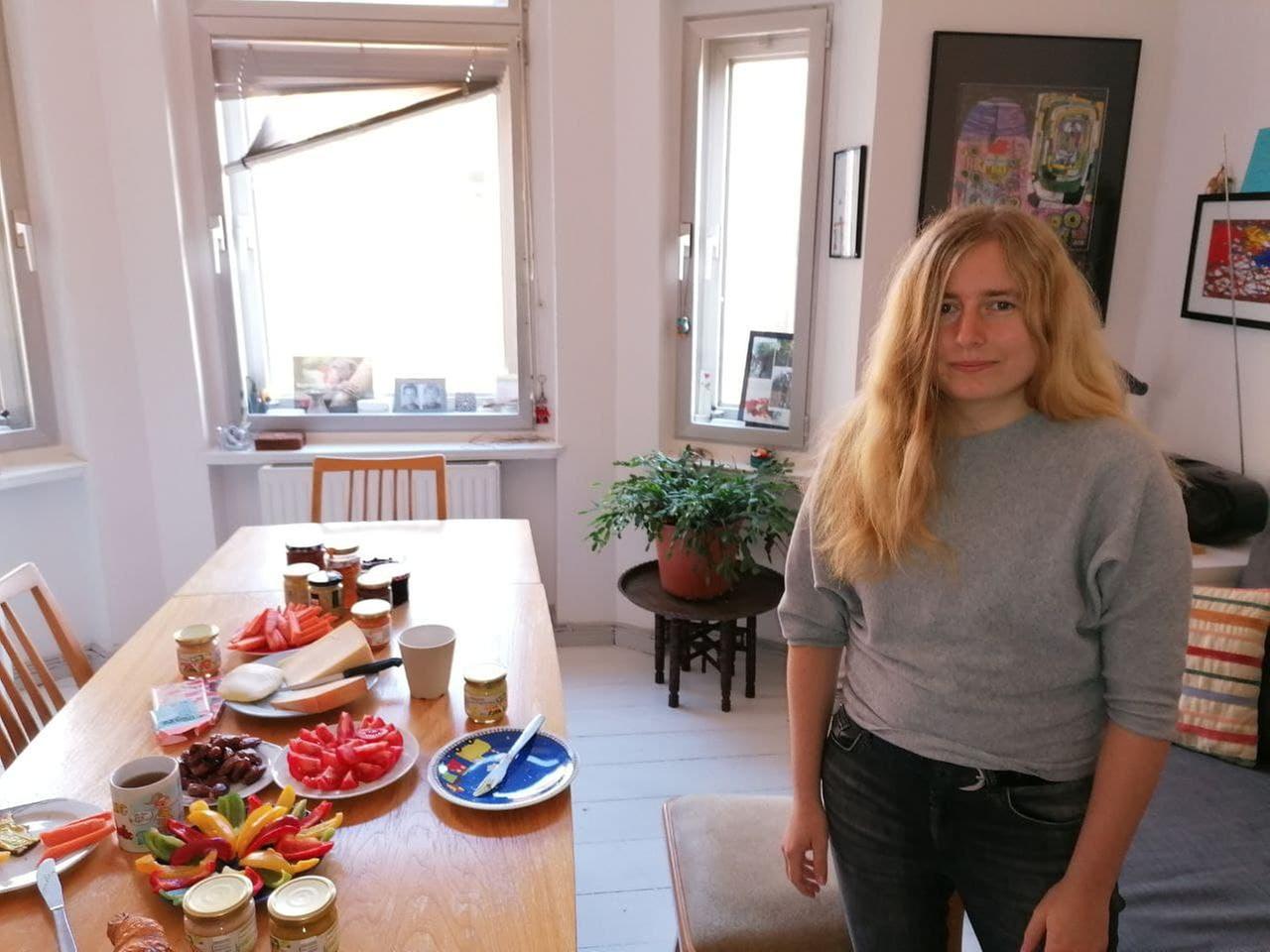 Eine junge Frau mit langen blonden Haaren in grauem Pullover und schwarzer Jeans steht neben einem mit Früchten, Paprikastreifen, Honig und Käse gedeckten Frühstückstisch.