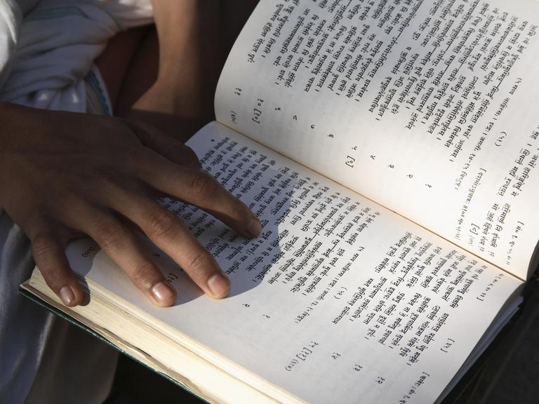 Eine Person hält ein offenes Buch in der Hand, in dem Verse auf Sanskrit stehen. Eine Person hält ein offenes Buch in der Hand, in dem Verse auf Sanskrit stehen.