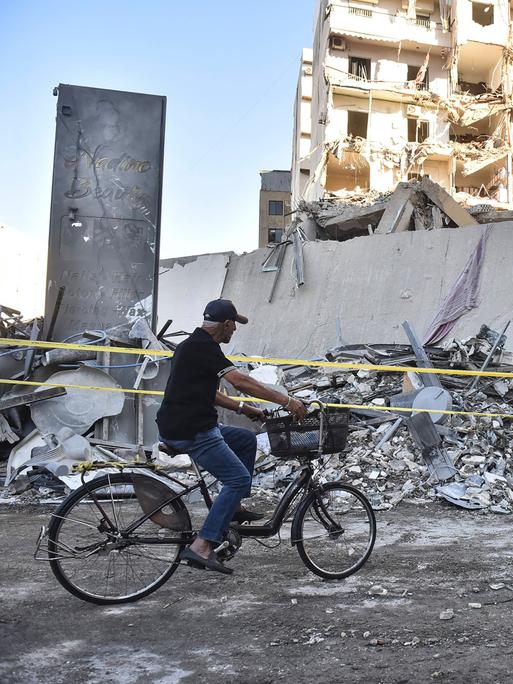 Ein Mann fährt auf einem Fahrrad vor einem völlig zerstörten Gebäude im Libanon vorbei.