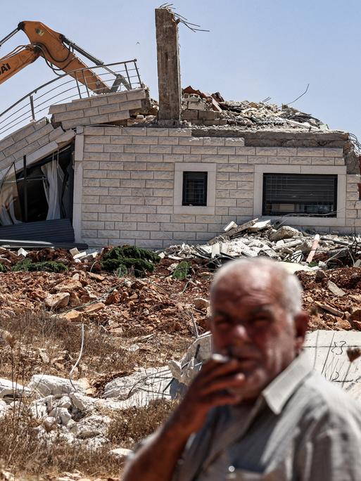 Ein Mann raucht eine Zigarette, während israelische Bagger am 4. August 2025 im Dorf Judeira südlich von Ramallah im besetzten Westjordanland ein Gebäude abreißen. 