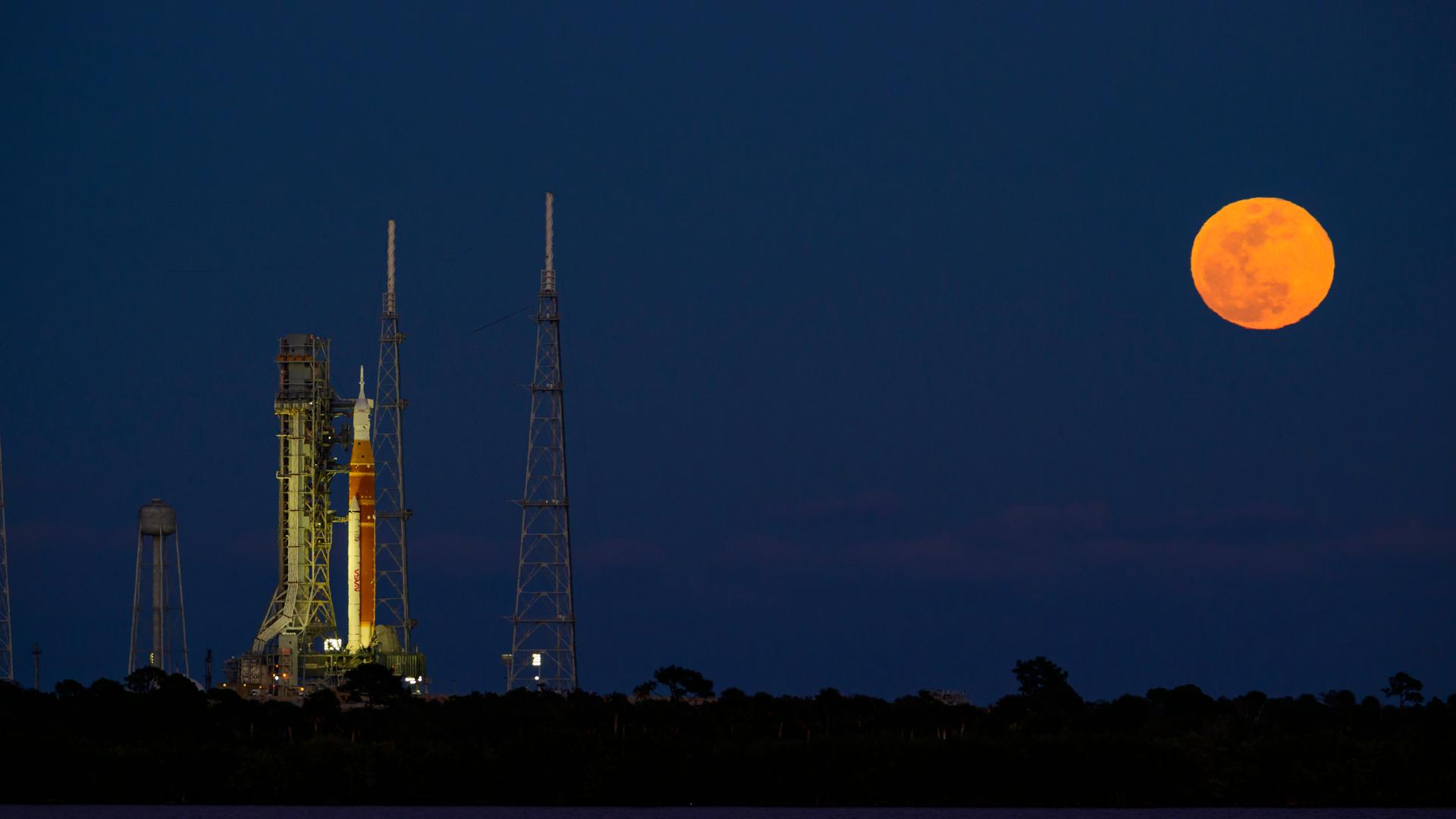 Nachthimmel am 01.02.2026 über Cape Canaveral in Florida. Links im Bild eine senkrecht stehende Orion-Rakete, rechts im Bild ein orangefarbener Vollmond.