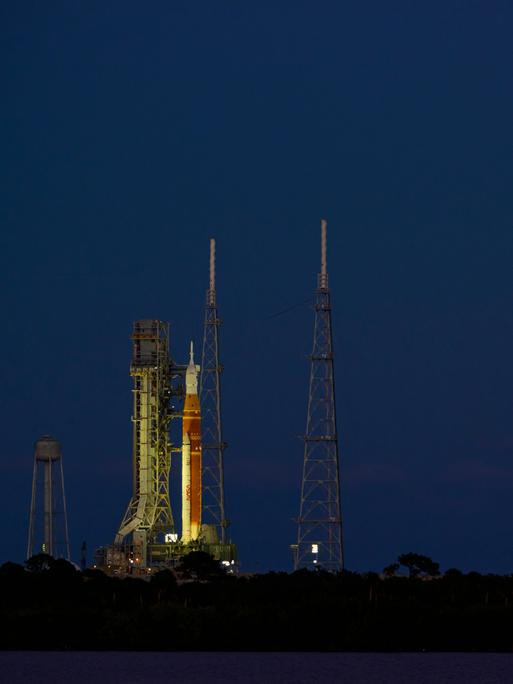 Nachthimmel am 01.02.2026 über Cape Canaveral in Florida. Links im Bild eine senkrecht stehende Orion-Rakete, rechts im Bild ein orangefarbener Vollmond.