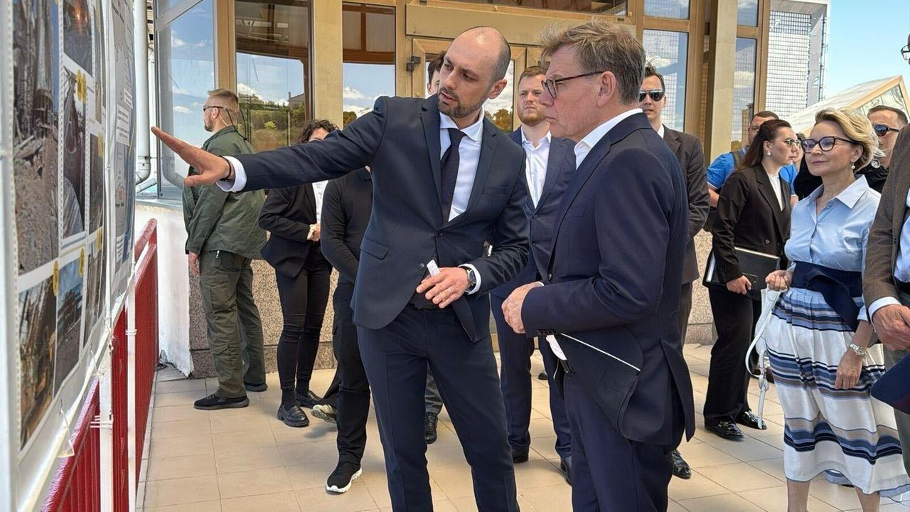 Wadephul (m.) vor einer Bilderwand. Ein Mann deutet auf ein Bild und scheint dem Minister etwas zu erläutern. Dahinter weitere Personen. Wadephul (m.) vor einer Bilderwand. Ein Mann deutet auf ein Bild und scheint dem Minister etwas zu erläutern. Dahinter weitere Personen.
