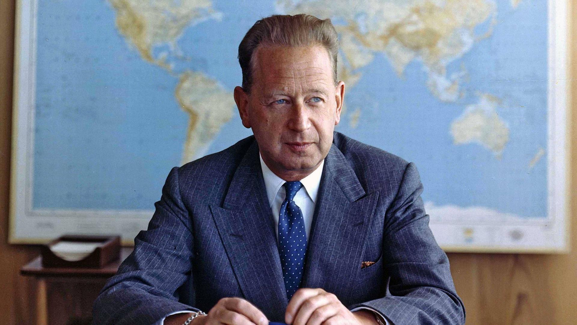 Dag Hjalmar Hammarskjöld sitzt in Anzug und Krawatte an einem Schreibtisch. Er guckt leicht nach links. In den Händen hält er einen Stift. Hinter ihm hängt eine Weltkarte.