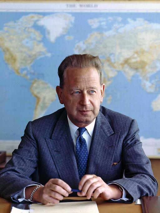 Dag Hjalmar Hammarskjöld sitzt in Anzug und Krawatte an einem Schreibtisch. Er guckt leicht nach links. In den Händen hält er einen Stift. Hinter ihm hängt eine Weltkarte.