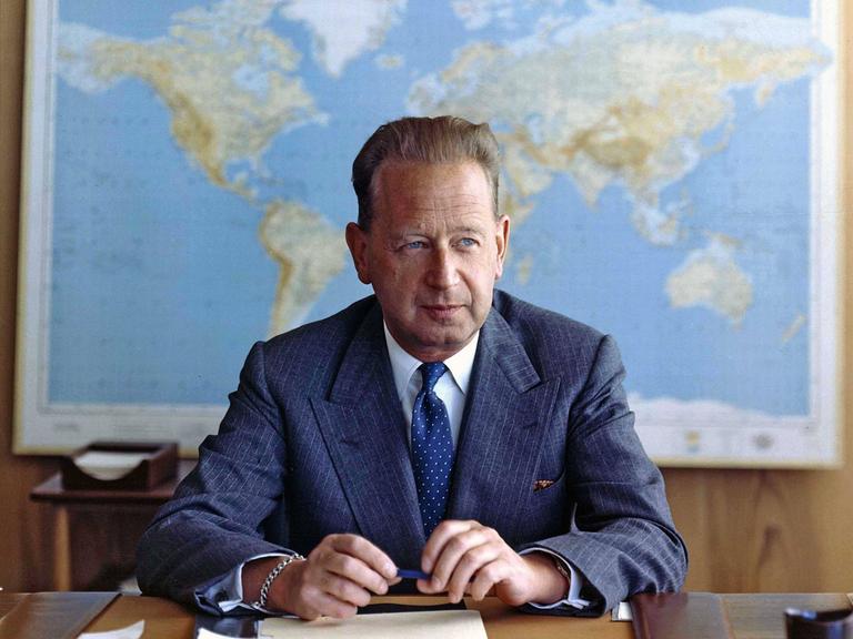 Dag Hjalmar Hammarskjöld sitzt in Anzug und Krawatte an einem Schreibtisch. Er guckt leicht nach links. In den Händen hält er einen Stift. Hinter ihm hängt eine Weltkarte.
