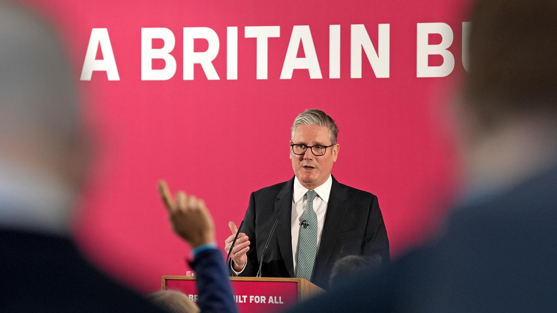 Der britische Premierminister Sir Keir Starmer hält eine Rede. Im Vordergrund sind Personen verschwommen von hinten abgebildet.