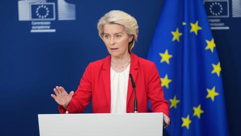 Ursula von der Leyen steht an einem Pult und spricht. Sie hat ihre rechte Hand leicht erhoben. Im Hintergrund ist eine EU-Flagge