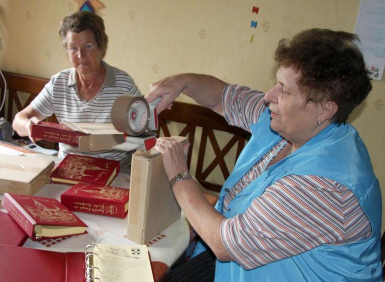 Drei ältere Damen sitzen an einem Tisch und packen Ringbücher in Pakete.