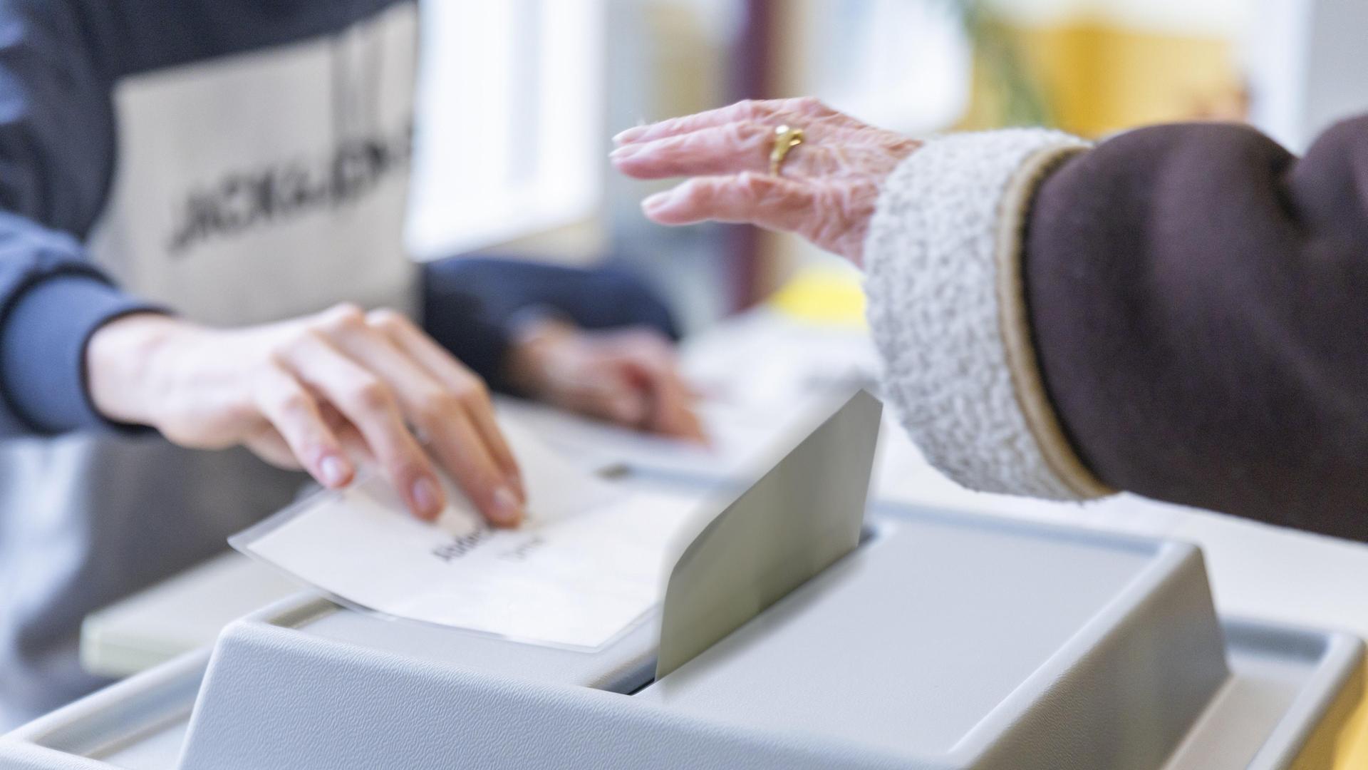 Stimmabgabe in einem Wahllokal bei der Bundestagswahl: Eine Hand wirft den Wahlschein in die Urne, die von einer anderen Hand freigegeben wird.